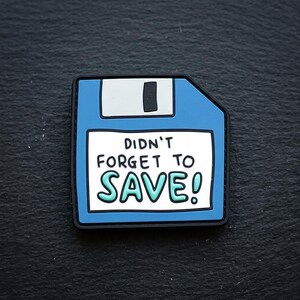 May include: A blue floppy disk pin with a white label featuring the black text DIDN'T FORGET TO and the teal text SAVE! The pin has a black border and a white sliding tab detail at the top, designed to resemble a classic computer storage device.