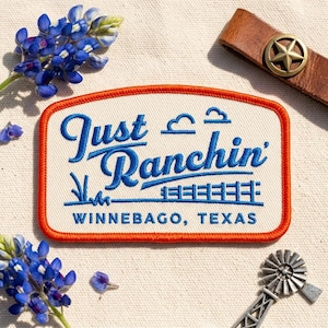 May include: An embroidered patch with the words "Just Ranchin' Winnebago, Texas" in blue script and block letters, framed by an orange border. The patch is surrounded by bluebonnet flowers, a leather strap with a star-shaped buckle, and a metal windmill.