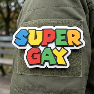 May include: A rectangular morale patch featuring the text SUPER GAY in a bold, multi-colored font style. The letters are blue, yellow, red, and green with black outlines, set against a white background. The patch is attached to an olive green fabric.
