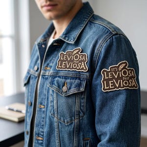 May include: A blue denim jacket featuring two identical rectangular patches with brown backgrounds and cream-colored borders. Each patch displays the text "IT'S LEVIOSA NOT LEVIOSA" in a stylized font, accompanied by a small feather graphic above the text.