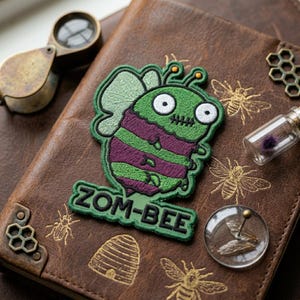 May include: Embroidered patch featuring a green and purple zombie bee with the text "ZOM-BEE". The patch is on a brown leather journal with bee-themed embellishments and a magnifying glass.