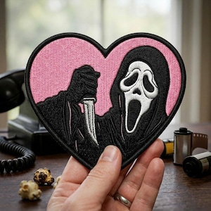 May include: A heart-shaped embroidered patch featuring a black-hooded figure with a white ghost mask holding a silver knife. The background of the patch is a solid light pink, and the entire design is outlined with a thick black border.