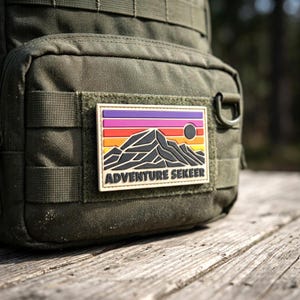 May include: An olive green tactical backpack featuring a rectangular morale patch. The patch displays a stylized mountain landscape with horizontal stripes of purple, pink, orange, and yellow, a black sun, and the text ADVENTURE SEKEER in black.