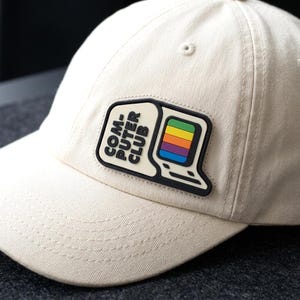 May include: A cream-colored baseball cap featuring a black and cream rubberized patch. The patch displays the text "COM-PUTER CLUB" in black lettering next to a stylized computer monitor icon with a multi-colored horizontal striped screen.