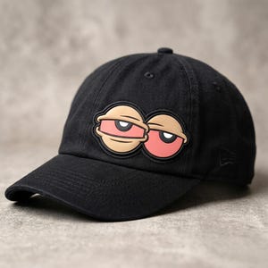 May include: A black baseball cap featuring a prominent rubberized patch on the front. The patch displays two cartoonish, round eyes with heavy, drooping eyelids, light tan skin tones, and large, circular, reddish-pink irises with black pupils.