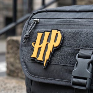 May include: A dark gray tactical bag featuring a yellow and black rubber patch with the letters HP in a stylized font. The bag includes a zippered compartment, a hook-and-loop panel, and a black plastic side-release buckle on the front flap.