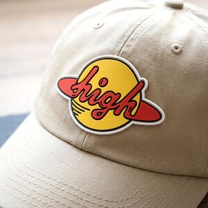 May include: A beige baseball cap featuring a circular patch on the front. The patch displays a yellow planet with a red ring and the word high written in red cursive script across the center. The cap has a structured crown and a curved brim.