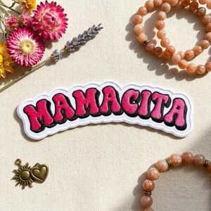 May include: A white patch with the word "MAMACITA" in pink bubble letters outlined in black. The patch is surrounded by dried flowers, sunstone bracelets, and a small charm.