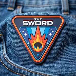 May include: A triangular rubber patch featuring the text THE SWORD in white letters. The design includes a central orange and yellow flame graphic against a dark blue background with light blue rays and white star accents, bordered by orange lines.