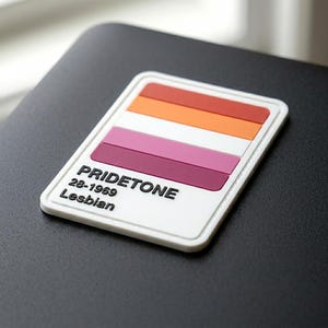 May include: A rectangular patch with a white border and background featuring a horizontal color block design in shades of red, orange, white, and pink. The text PRIDETONE, 28-1969, and Lesbian is printed in black on the lower left side.