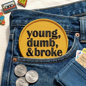 May include: A pair of blue denim jeans with a yellow circular patch in the pocket that reads "young, dumb, & broke" in black lettering. Three quarters are visible, along with a wallet and some pins.