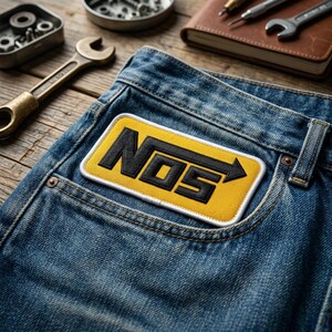 May include: A rectangular embroidered patch featuring the black text NOS with an arrow pointing right, set against a yellow background with a white border, attached to the front pocket area of a pair of blue denim jeans with visible stitching.