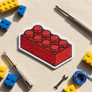 May include: A red embroidered patch in the shape of a classic building block, with a white border. The patch is surrounded by yellow and blue toy blocks, screwdrivers, a small wrench, and a ball of blue string, all on a neutral background.