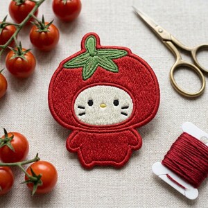 May include: An embroidered patch featuring a white cat character wearing a red tomato costume with a green stem top. The patch is surrounded by fresh red cherry tomatoes, a pair of gold-toned metal scissors, and a spool of red embroidery thread.