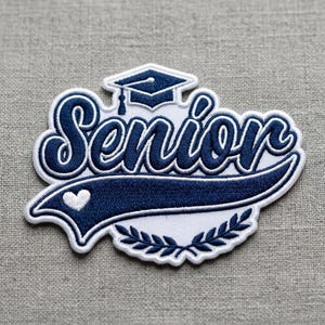 May include: An embroidered patch featuring the word Senior in dark blue script lettering. Above the text is a dark blue graduation cap with a tassel. Below the text is a dark blue swooping banner with a small white heart and a laurel wreath.