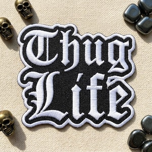 May include: Black and white embroidered patch with the words "Thug Life" in a gothic font. The patch has a black background and a white border. Decorative skulls and stones are scattered around the patch.