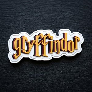 May include: A die-cut patch featuring the word "gryffindor" in a stylized, layered font. The lettering is primarily yellow with dark red and black shadow accents, set against a white background with a stitched border outline.
