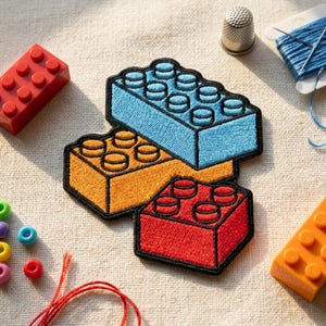May include: Embroidered patches in the shape of toy building blocks in blue, orange, and red. The patches are on a neutral fabric surface, with a red building block and an orange building block visible. Sewing notions are also in the image.