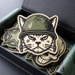 May include: A rubberized morale patch featuring a stylized cat wearing an olive green military helmet. The cat has a stern expression with black line art details on a cream-colored background. The patch is shaped like the cat's head and ears.