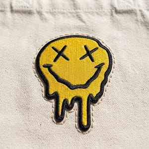May include: Embroidered patch featuring a yellow, melting smiley face with black outlines and crossed-out eyes. The patch is sewn onto a beige fabric, creating a contrast between the bright yellow and the neutral background. The design has a distressed, edgy aesthetic.