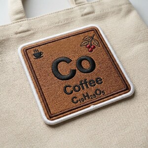 May include: A beige canvas tote bag featuring a square embroidered patch. The patch is brown with a white border and displays the black text Co, Coffee, and C19H28O5, along with a small black coffee cup icon and a cluster of red coffee cherries.