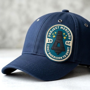 May include: A navy blue baseball cap featuring a rectangular patch with rounded corners. The patch displays a graphic of a space capsule, yellow stars, and the text "PROJECT MERCURY 1962 FIRST AMERICAN TO ORBIT" in a cream and teal color scheme.