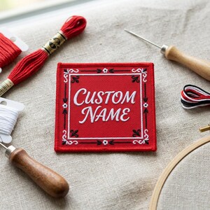 May include: A red embroidered patch with the words "CUSTOM NAME" in white script. The patch has a decorative black and white border. Embroidery floss, needles, and a small embroidery hoop are arranged around the patch.