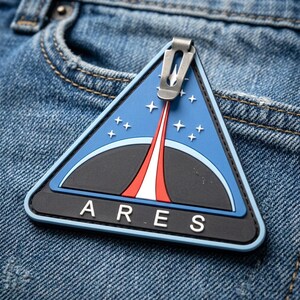 May include: A triangular rubber patch featuring a blue background with a black planet silhouette, a red and white rocket trail, and white stars. The word ARES is printed in white capital letters at the bottom, and a metal clip is attached to the top.