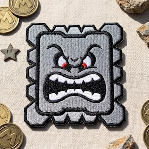 May include: Embroidered patch featuring a gray Thwomp character from the Super Mario video game series. The patch has a square shape with a jagged border, a black mouth, and red eyes. The patch is surrounded by gold coins and a star.