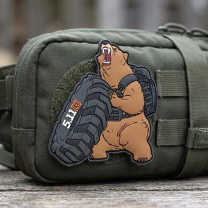May include: An olive green tactical pouch featuring a rubberized morale patch. The patch depicts a brown bear wearing a tactical vest and carrying a large black tire. The text 5.11 is printed in white on the tire with an orange square logo.