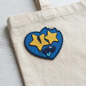 May include: A beige canvas tote bag with a blue heart-shaped patch. The patch features two yellow stars, a blue tongue, and a bite taken out of the top. The patch is embroidered with a white outline.