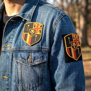 May include: A blue denim jacket featuring two shield-shaped patches. Each patch displays a black, red, and yellow design with a Union Jack in the corner and a central emblem featuring a stylized flower. The patches have a yellow border.
