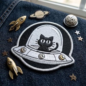 May include: A large embroidered patch featuring a black cat inside a silver UFO, surrounded by two brass-toned rocket pins, a planet patch, a moon-textured dome, and several small metallic star-shaped studs on a dark blue denim fabric background.
