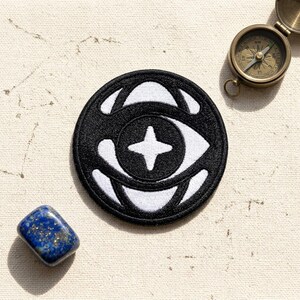 May include: A black and white embroidered patch with an eye design and a compass. The patch features a circular shape with a stylized eye and a four-pointed star in the center. A small, polished blue stone is also present.