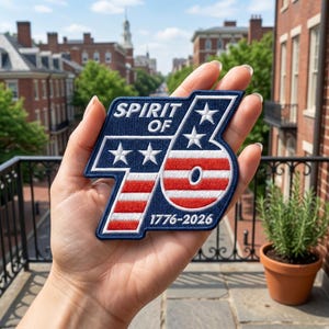 May include: A patriotic patch with the text "Spirit of 76" and the years 1776-2026. The patch features a blue background with white stars and red and white stripes, resembling the American flag. The patch is held up in front of a blurred background.