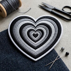 May include: A heart-shaped embroidered patch featuring concentric rings of black, dark gray, medium gray, and light gray thread. The patch has a thick white stitched border and is displayed alongside a spool of gray thread, black scissors, and two pins.