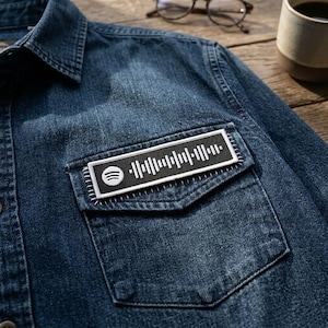 May include: A dark blue denim shirt with a chest pocket featuring a black and white Spotify patch. The patch displays the Spotify logo and a barcode-like design. The shirt has a classic collar and button-down front.