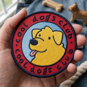 May include: A circular embroidered patch featuring a yellow dog with a black nose and pink tongue against a light blue background. The design is enclosed by a red border with the dark blue text "cool dogs club" stitched around the perimeter.