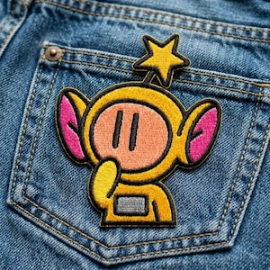 May include: A yellow embroidered patch featuring a cartoon character with a star on its head, pink ears, and a peach-colored face. The patch is sewn onto a blue denim pocket.