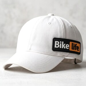 May include: A white baseball cap featuring a rectangular black and orange rubberized patch on the side. The patch displays the text "Bike life" in white and black lettering. The cap has a structured crown, a curved brim, and stitched eyelets.