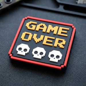 May include: A square morale patch with a black background and a red border. The text GAME OVER is displayed in a yellow pixelated font. Below the text are three white pixelated skull icons arranged horizontally across the center of the patch.