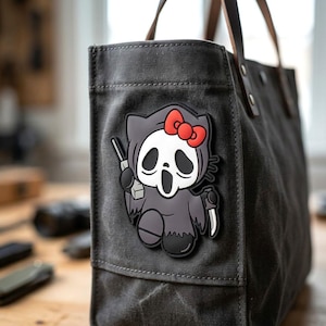 May include: A dark gray canvas tote bag featuring a rubberized patch of a cartoon character wearing a ghost mask, a black hooded robe, and a red bow. The character holds a silver knife in one hand and is depicted in a stylized, graphic design.