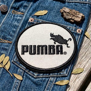 May include: A denim jacket with a circular patch featuring the word "PUMBA" in black capital letters. The patch has a black border and a black illustration of a boar. The jacket is on a wooden surface with leaves.