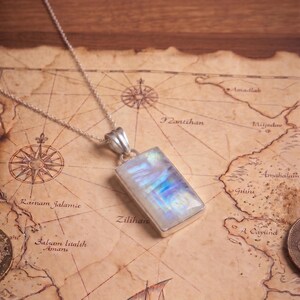 May include: A rectangular moonstone pendant set in a silver bezel, suspended from a delicate silver chain. The gemstone displays a translucent, iridescent sheen with flashes of blue and white light, resting against a vintage-style map background.
