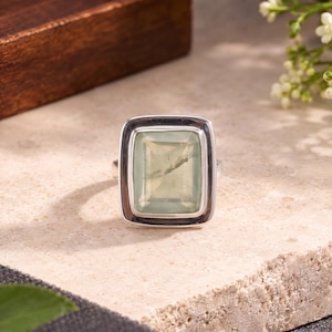 May include: A silver ring featuring a large, rectangular, faceted pale green gemstone set in a smooth, polished metal bezel. The stone has a translucent appearance with subtle internal inclusions, framed by a thick, rounded silver border.