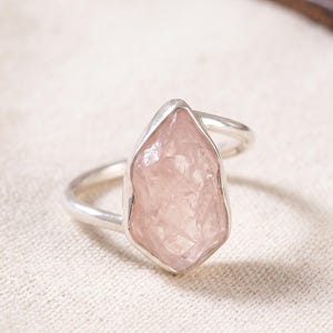 Op de afbeelding: Een zilveren ring met een ruwe, ongeslepen, lichtroze rozenkwarts edelsteen gezet in een gladde metalen zetting. De steen heeft een gefacetteerde, onregelmatige oppervlaktestructuur en is gemonteerd op een eenvoudige, gepolijste zilveren ring.