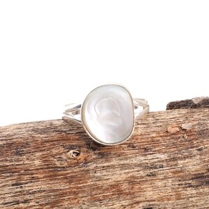 Könnte beinhalten: Ein Silberring mit einem großen, ovalen, perlmuttfarbenen weißen Stein. Der Stein ist in einem schlichten, modernen Design mit geteiltem Band gefasst. Der Ring wird auf einem Stück verwittertem Holz präsentiert.