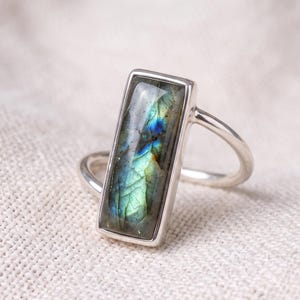 Puede incluir: Un anillo de plata con una piedra preciosa de labradorita rectangular de superficie lisa y pulida. La piedra muestra destellos iridiscentes de colores azul, verde y dorado dentro de su textura natural agrietada, montada en una banda de metal simple.