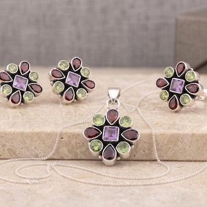 May include: A jewellery set consisting of a pendant on a silver chain, a pair of stud earrings, and a ring. Each piece features a central square-cut purple amethyst surrounded by alternating teardrop-shaped red garnets and round green peridot stones.
