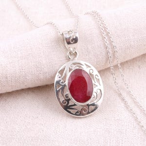 May include: A silver pendant necklace featuring an oval-shaped, deep red gemstone set in a decorative, openwork silver frame with swirling patterns. The pendant hangs from a delicate silver chain, showcasing a polished finish and intricate metalwork.